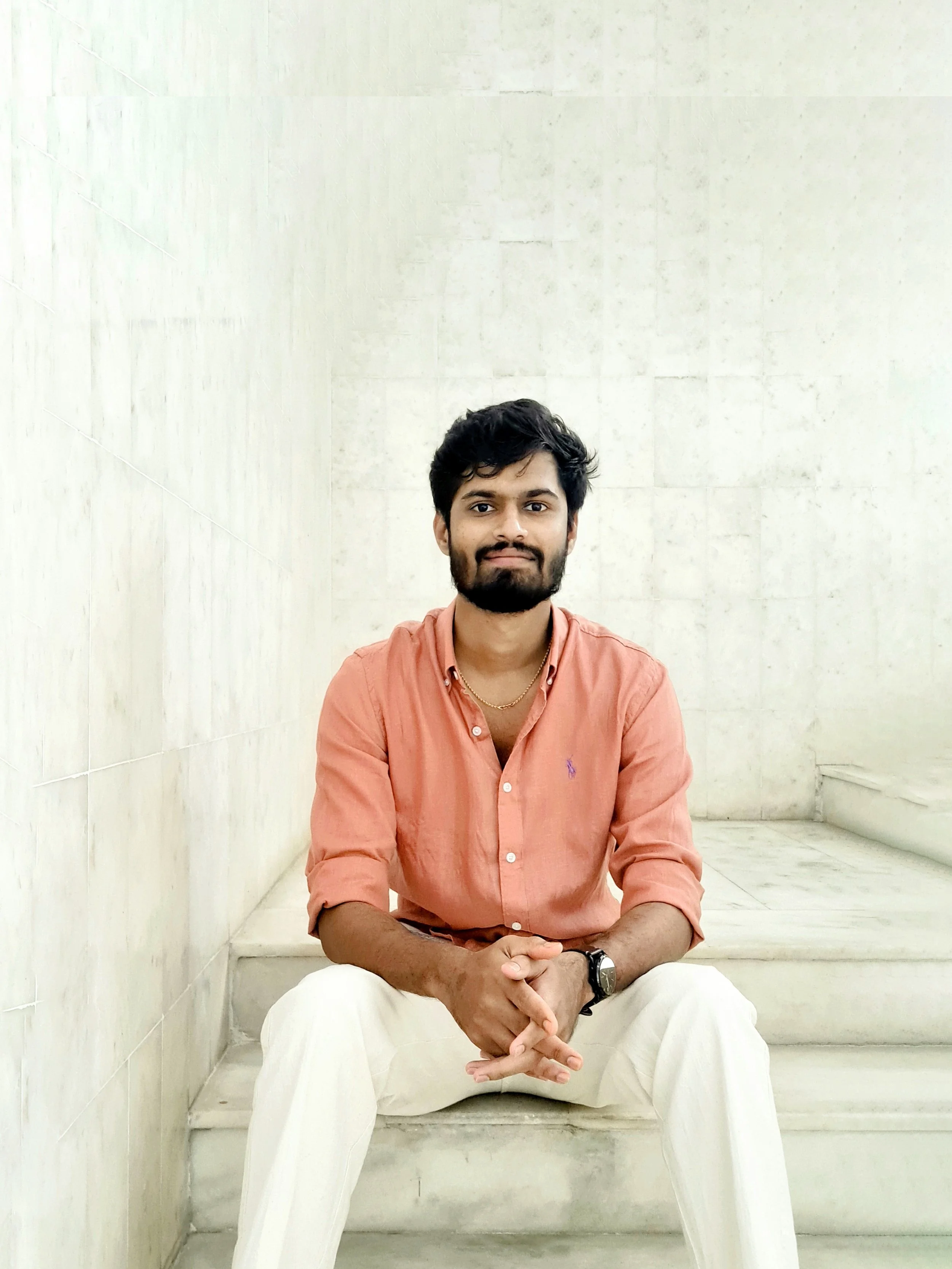 A young man with dark hair and a beard sitting on white marble stairs, wearing a peach-colored button-up shirt and white pants, with a black wristwatch and a gold chain, against a beige stone wall background.