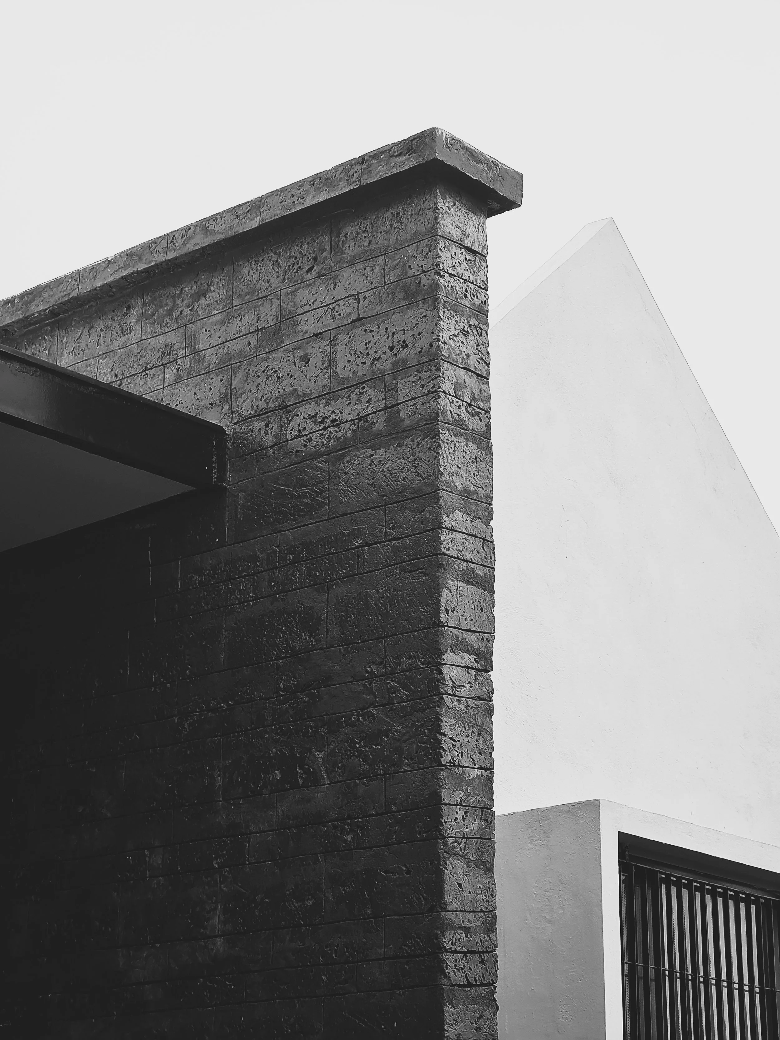 Close-up black and white photo of a building corner with stone and stucco walls, with a small window with bars.