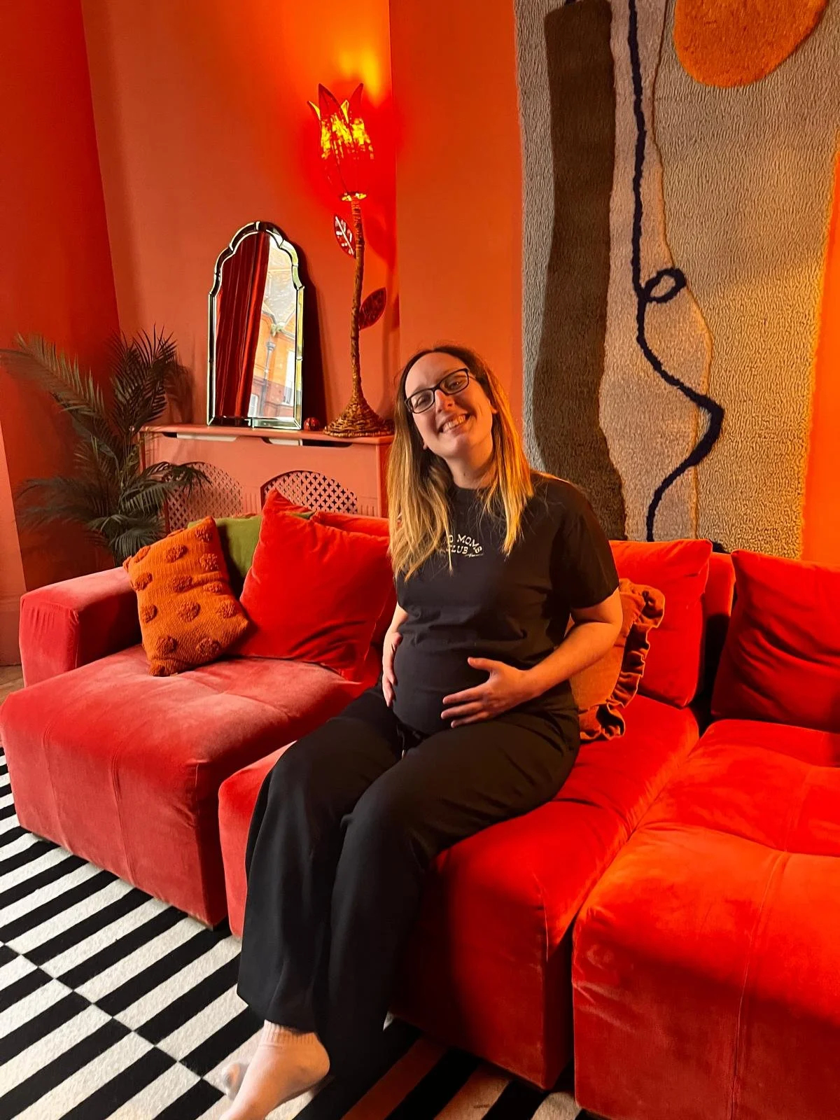 A woman with long hair and glasses sitting on a red sofa in a vividly decorated room. She is smiling, holding her pregnant belly, with orange cushions around her. The room has a dark striped rug, a tall lamp with an orange shade, a mirror with a wooden frame, and a tapestry on the wall.