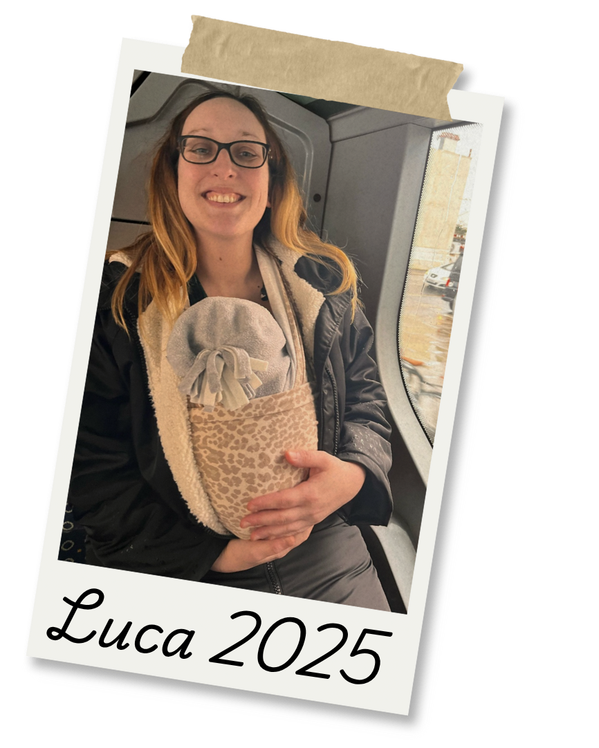 A woman with glasses and long hair, smiling, holding a small beige bag containing a gray plush toy inside a patterned pouch, sitting in a vehicle with a window showing a rainy parking lot outside. The photo has a Polaroid-style border with the handwritten text "Luca 2025" at the bottom.