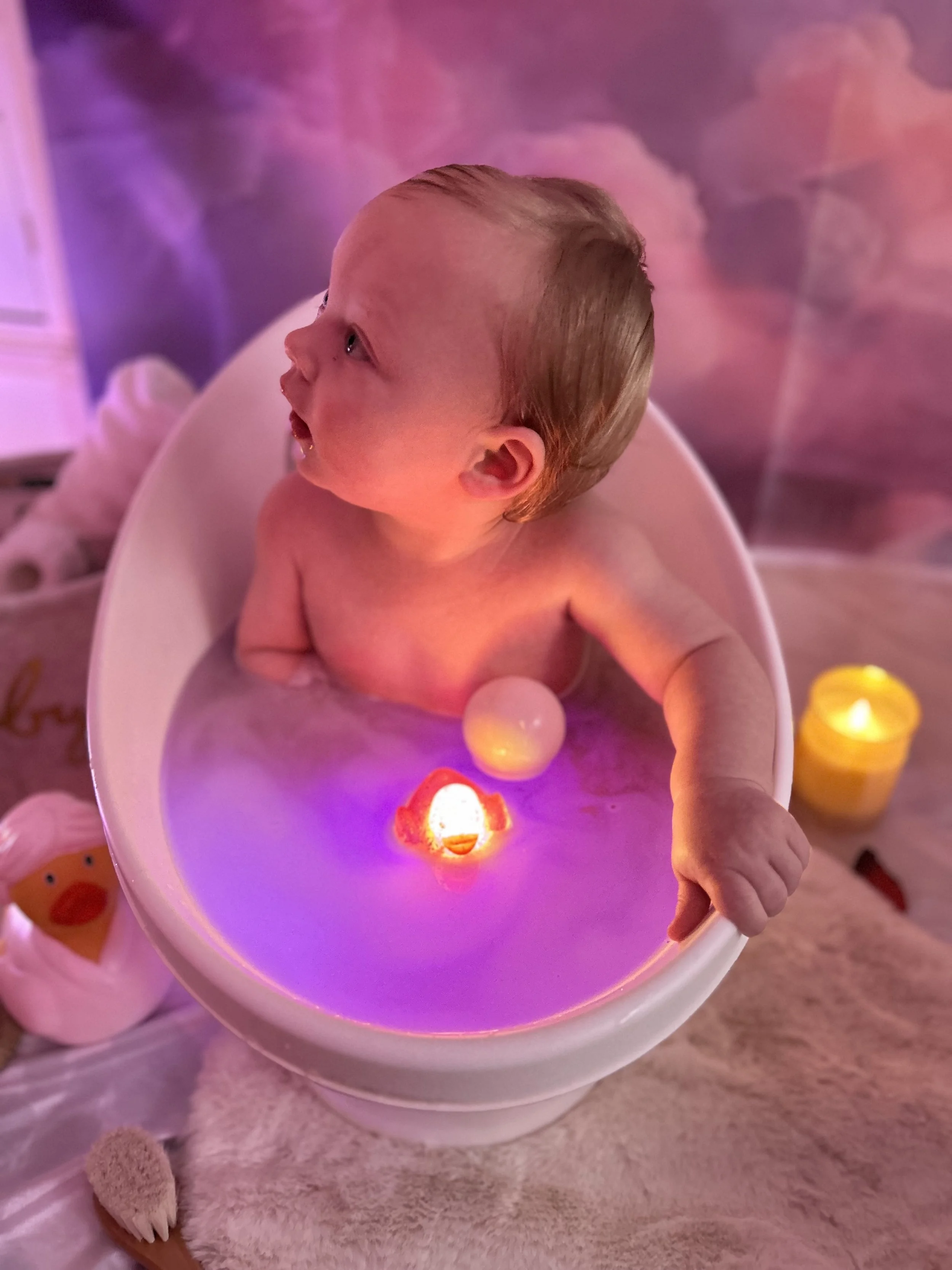 A young child with light brown hair sitting in a white bathtub filled with water, illuminated by pink and purple lights. The child appears to be looking up, drawing attention to the scene's glow. Nearby are a yellow candle, a toy resembling a duck, and a bath brush on a soft white surface.