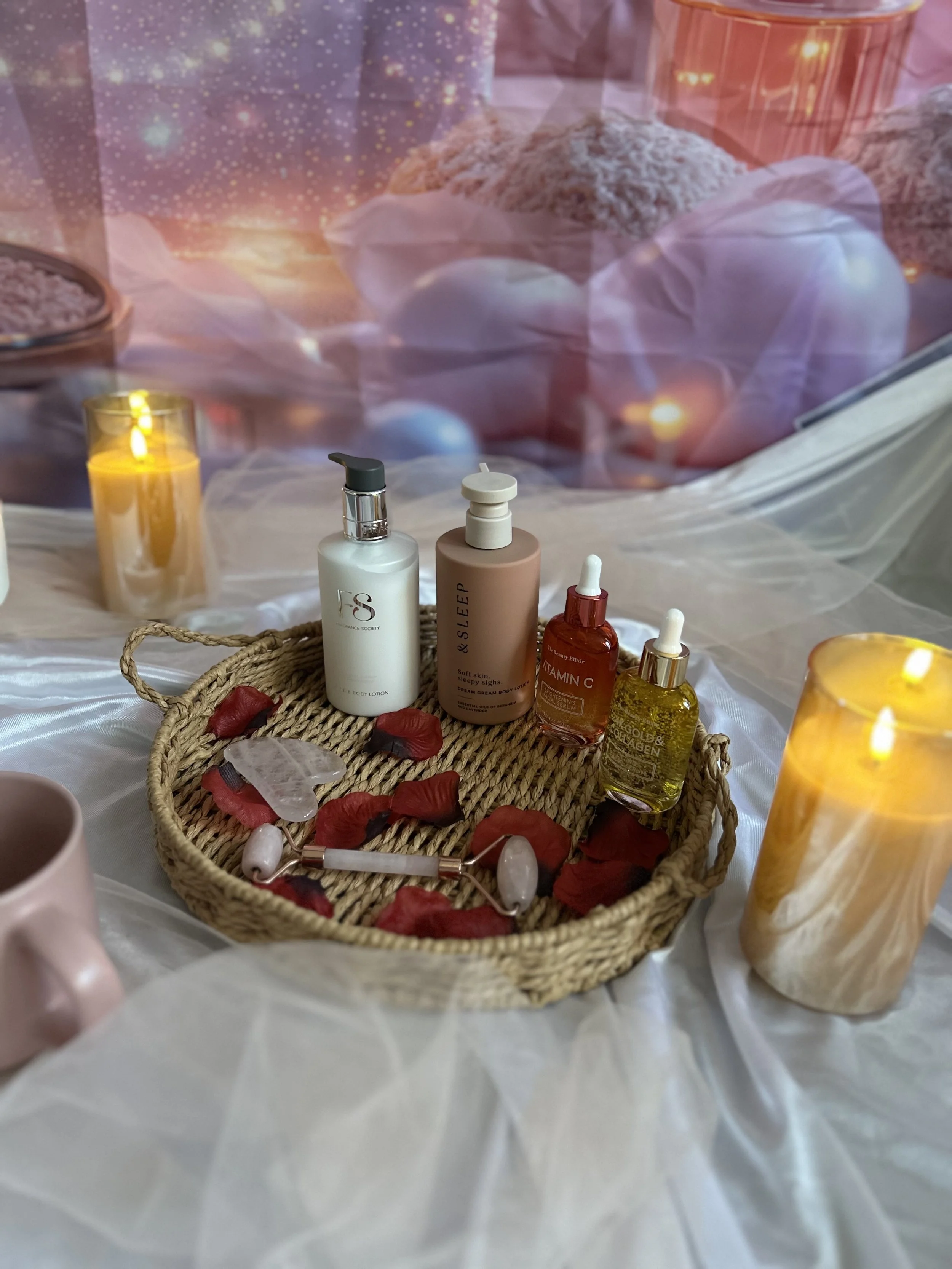 A wicker tray with skincare products and rose petals on a table, with candles and a pastel background.