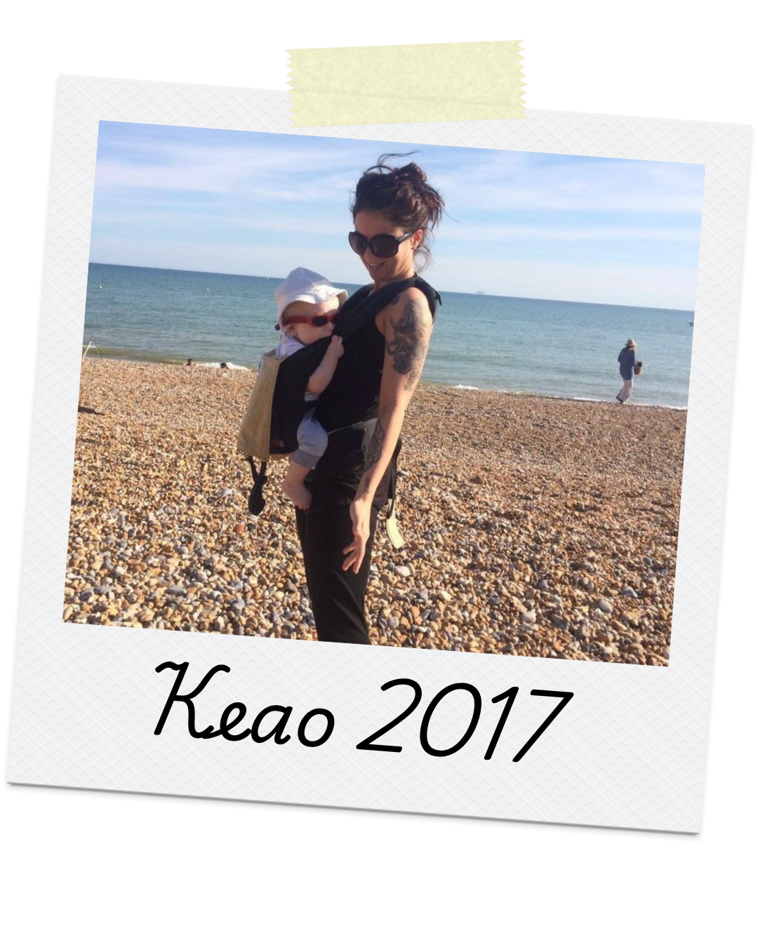 A woman with tattoos and sunglasses carrying a baby in a baby carrier on a rocky beach with the ocean and a person in the background. The photo has a Polaroid-style frame with yellow tape at the top and the text "Keao 2017" at the bottom.