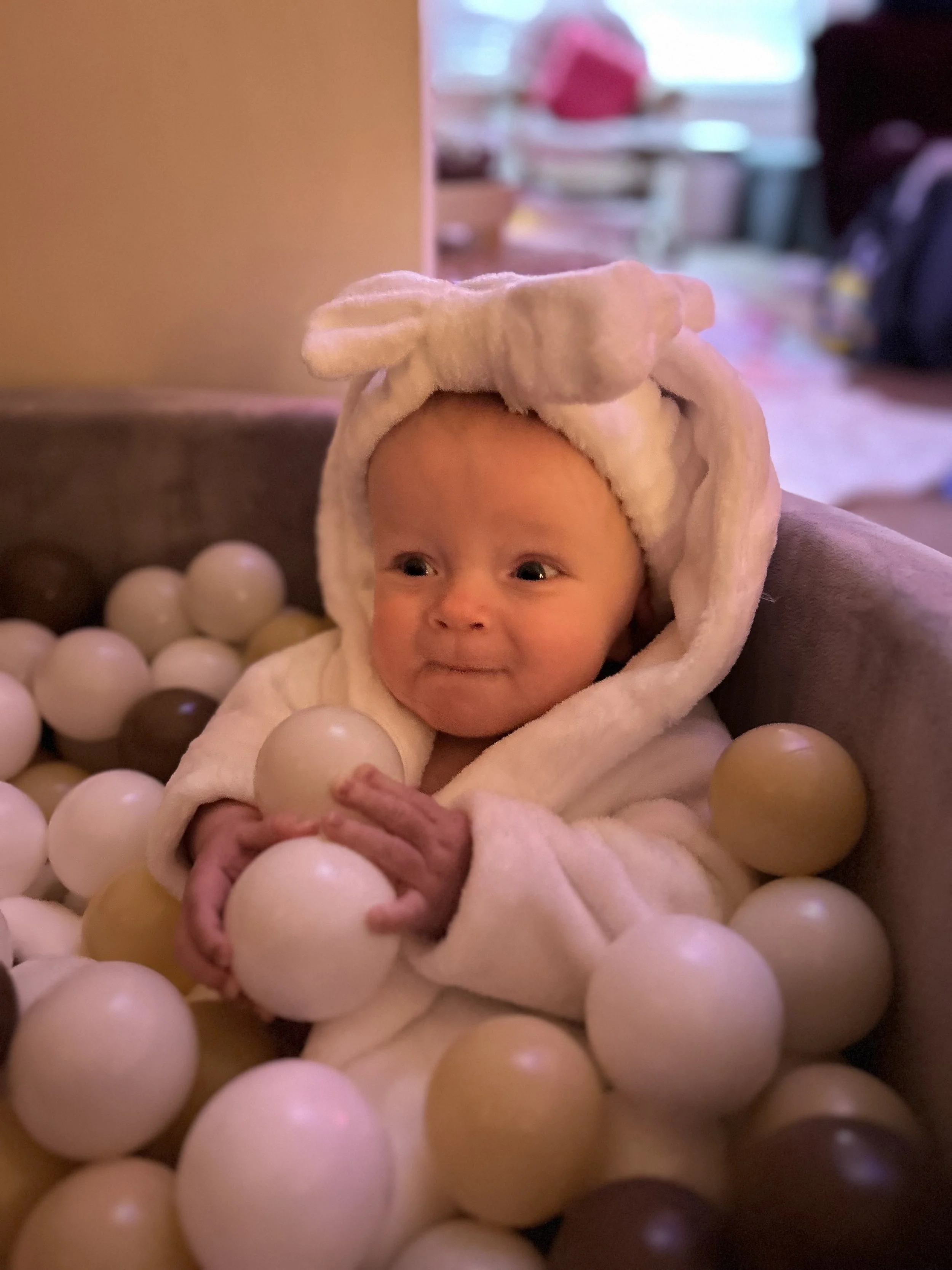 A baby wearing a hooded towel with bear ears sitting in a ball pit surrounded by cream, white, and brown plastic balls.