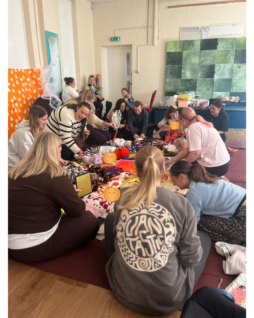 A group of adults and babies sitting on mats and a blanket on the floor, decorated with Halloween decorations like pumpkins, spider webs, and tinsel, in a room with a cream brick wall and a green tiled backdrop.