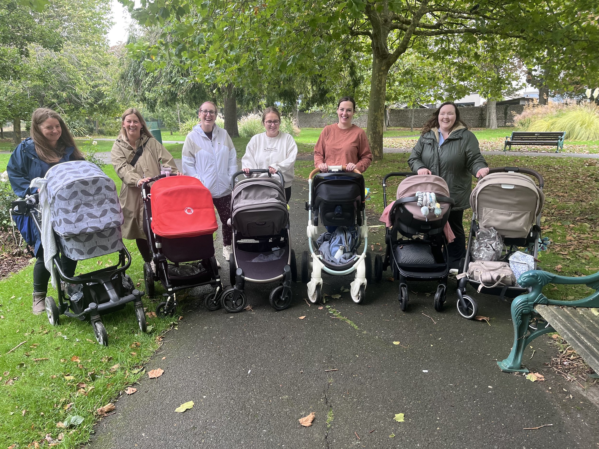 Group of seven women standing behind strollers in a park with trees and benches.