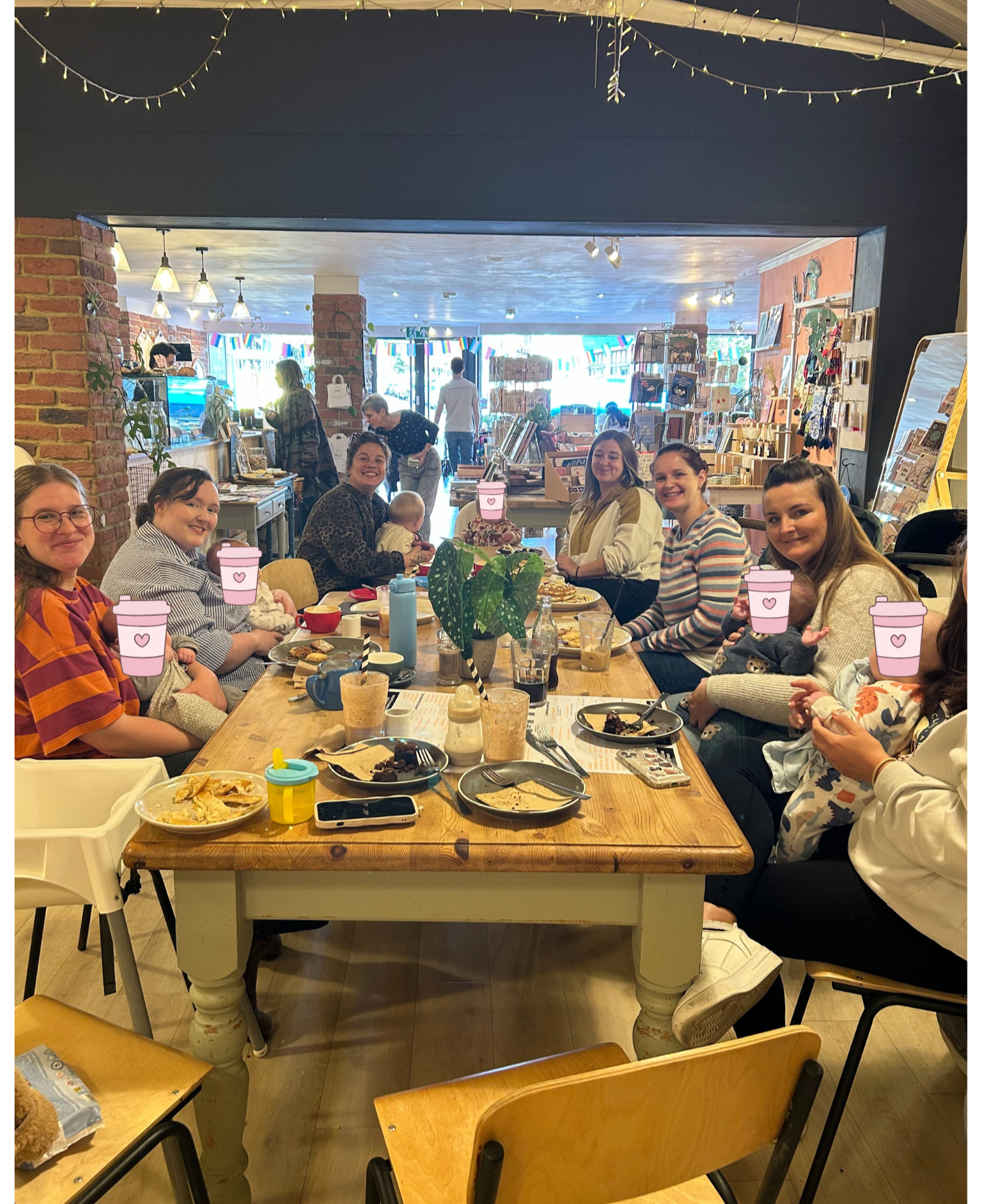 Group of women and children sitting around a wooden table in a cafe, with food and drinks, some women holding babies, all smiling, with a store behind them and fairy lights hanging from the ceiling.