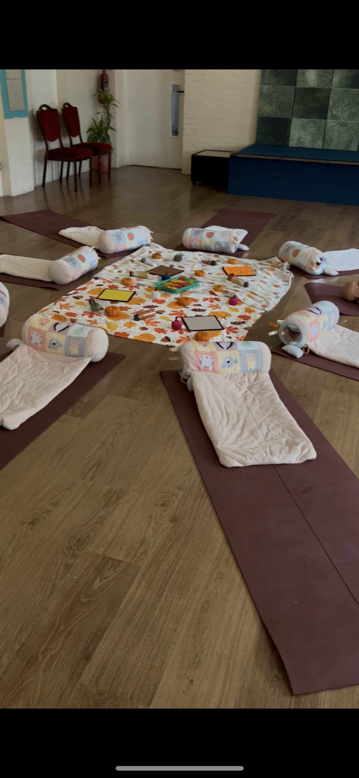 A children's yoga class setup with mats and rolls, pumpkins, toys, and a colorful fall-themed blanket in the center of the room.