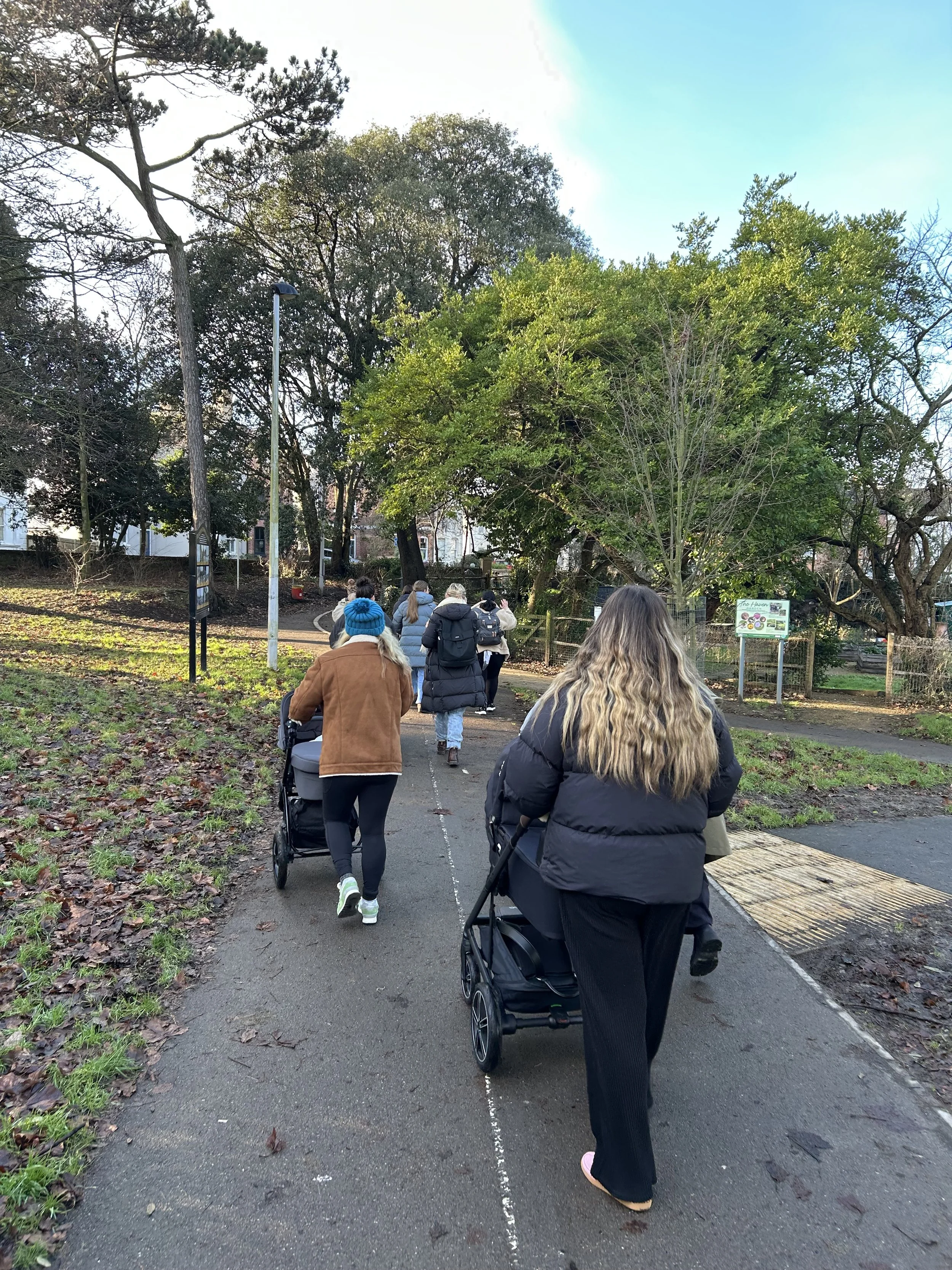 People walking on a paved path in a park, some pushing strollers, surrounded by trees and greenery, with clear sky above.