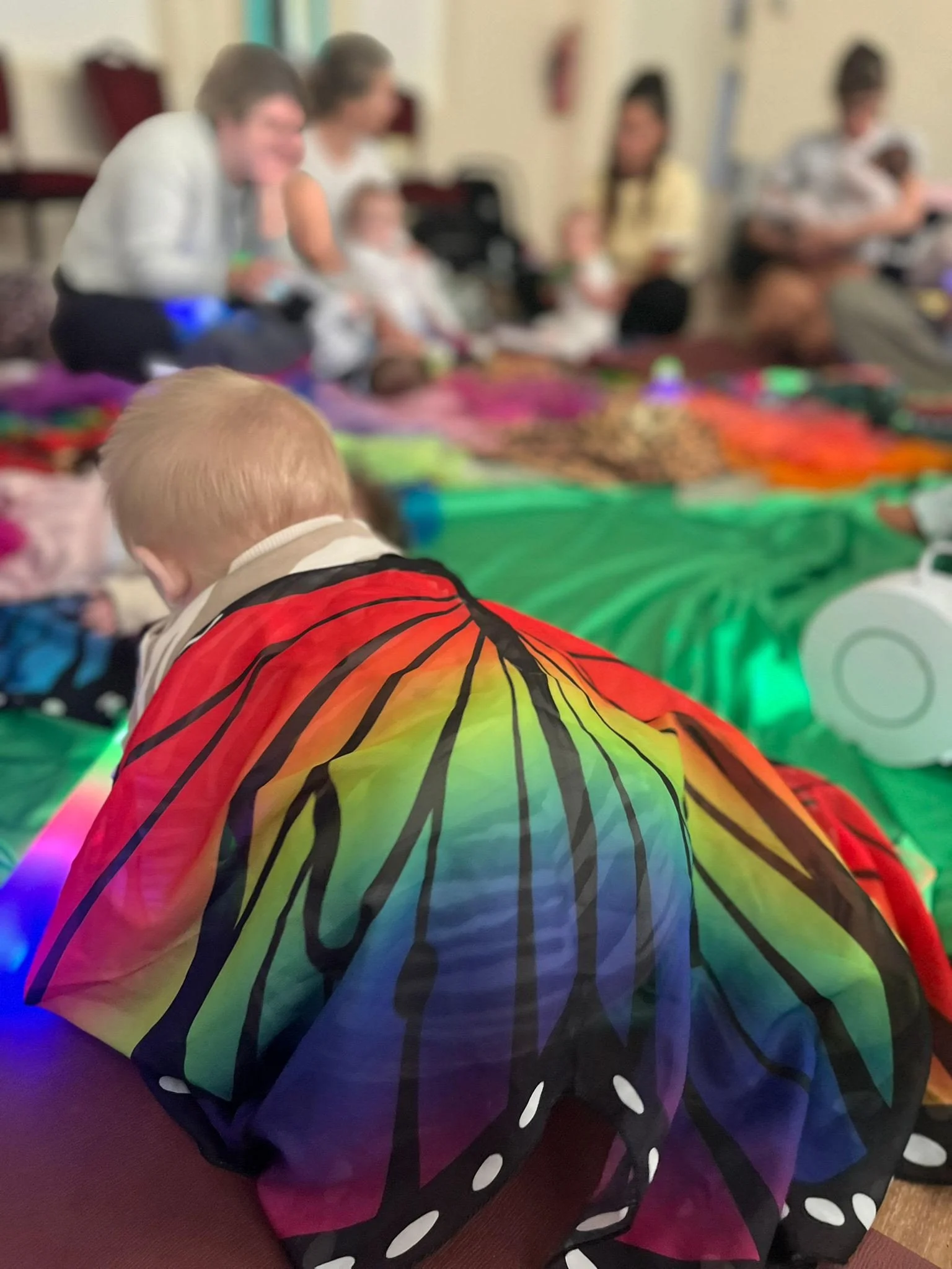 A group of people, including children, gathered around a table covered with colorful fabrics, with a child in the foreground wearing a rainbow-colored butterfly cape.