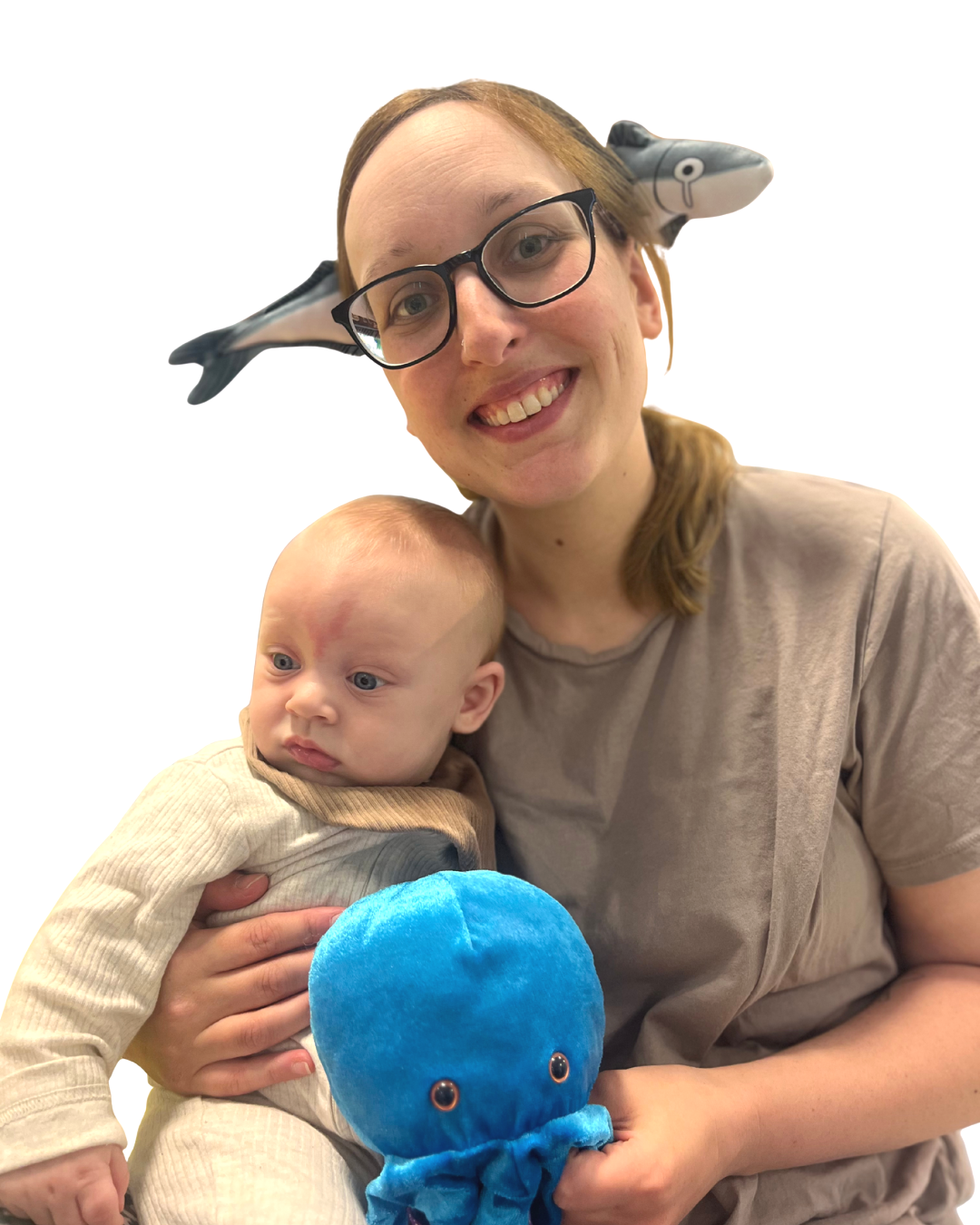 A woman with glasses and a dolphin-shaped headband holding a baby. The woman is smiling, and the baby has a serious expression. The woman is holding a blue plush octopus toy.