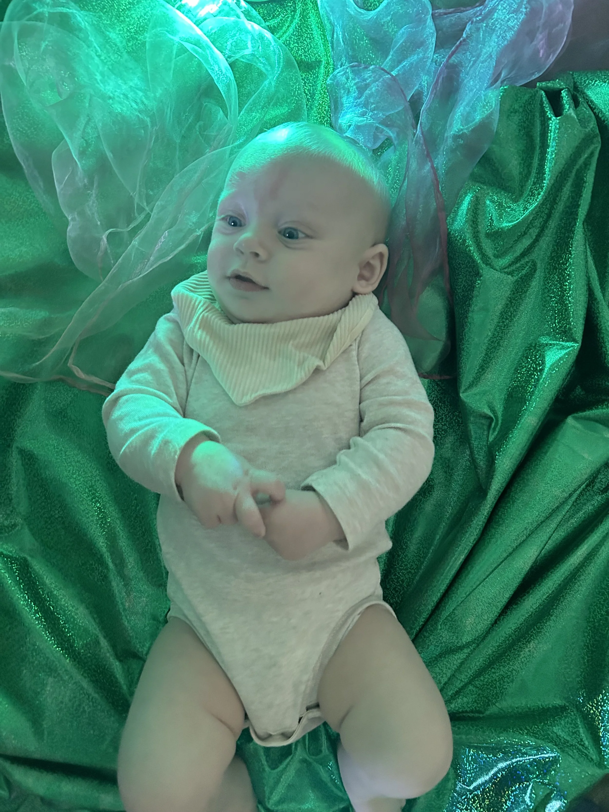A baby lying on a bed with green shimmering fabric and translucent green and purple fabric surrounding them.