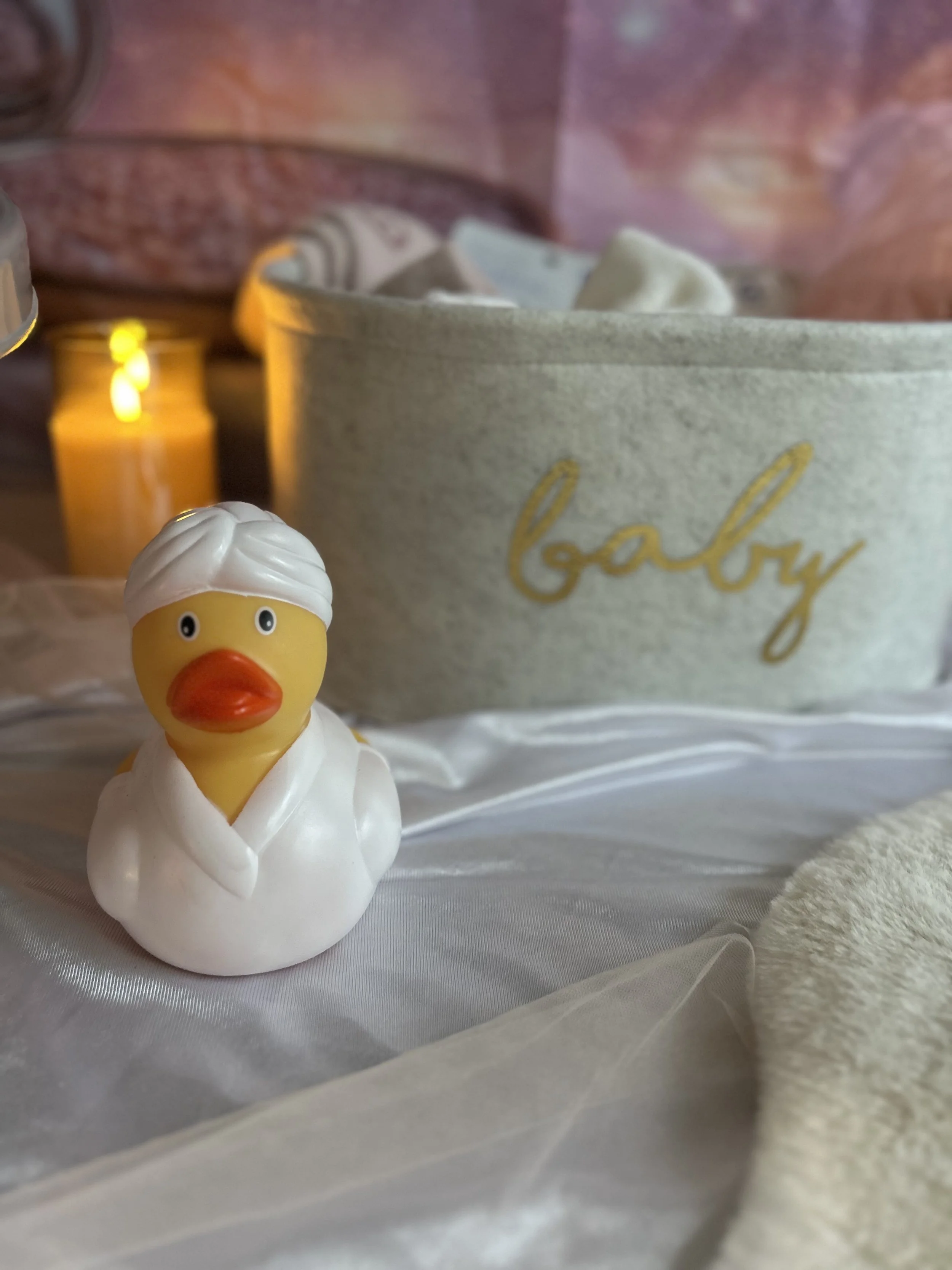A rubber duck dressed as a chef with a white chef's coat and white head covering, placed on a silky white fabric surface, with a large container of baby bath products labeled 'baby' and a lit yellow candle in the background.