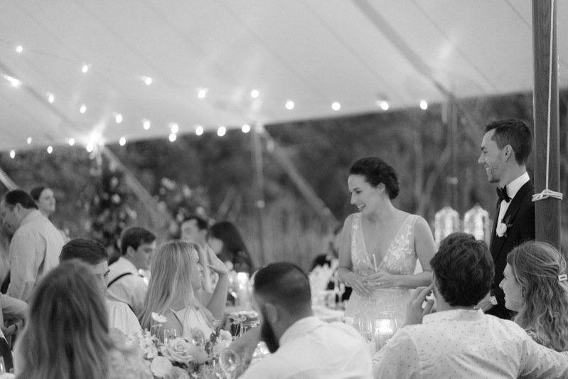 Group of people at a wedding reception under a tent with string lights, with a bride and groom standing and smiling at friends seated at a long table.