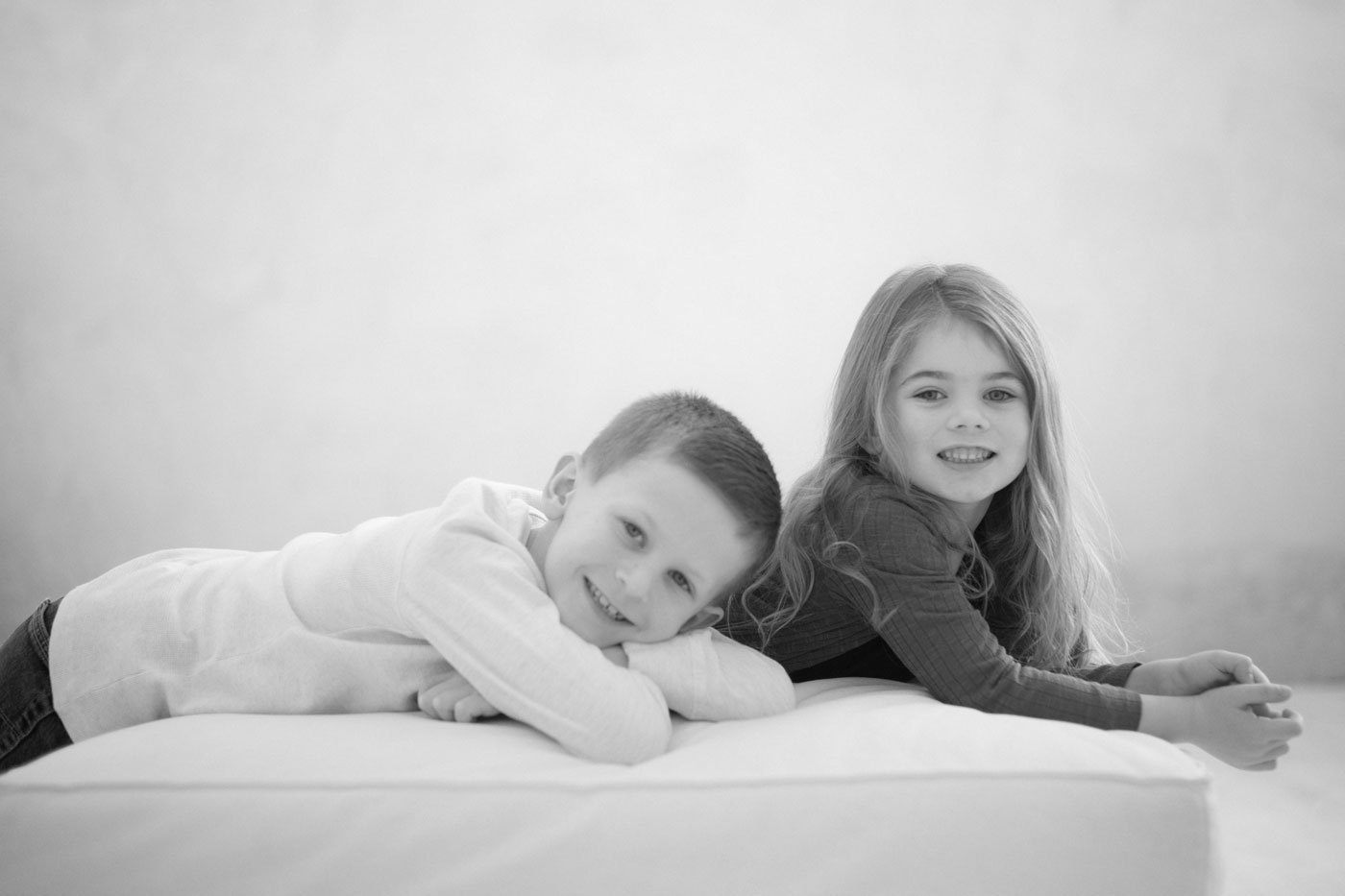 Black and white photo of a smiling boy and girl lying on a bed, facing the camera, in a minimal setting with a plain background.