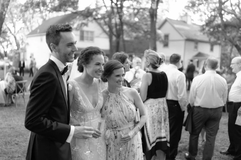 Group of people at an outdoor wedding reception, with a young couple in tuxedo and wedding dress smiling in the foreground.