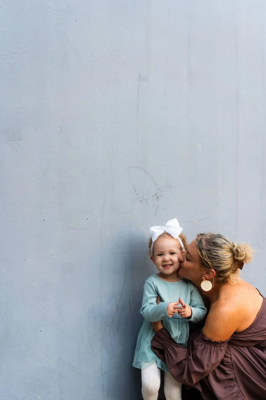A woman and a young girl share a kiss against a plain grey wall. The girl is smiling and wearing a light blue dress and white tights, with her hair in curly puffs and a large white bow. The woman is dressed in a mauve off-shoulder top with her hair i