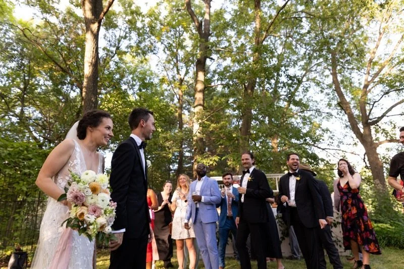 A bride and groom are standing outdoors, surrounded by wedding guests. The bride is holding a bouquet and wearing a white wedding dress. The groom is in a black tuxedo. Guests are smiling and chatting in a wooded setting with tall trees.