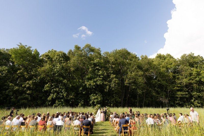 Outdoor wedding ceremony with guests seated on wooden chairs, officiant, bride and groom standing under a tree-lined sky.