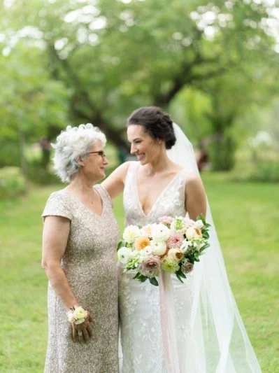Bride in a white wedding dress holding a bouquet, smiling and talking to an older woman in a gold dress with gray curly hair, outside on a grassy area with trees in the background.