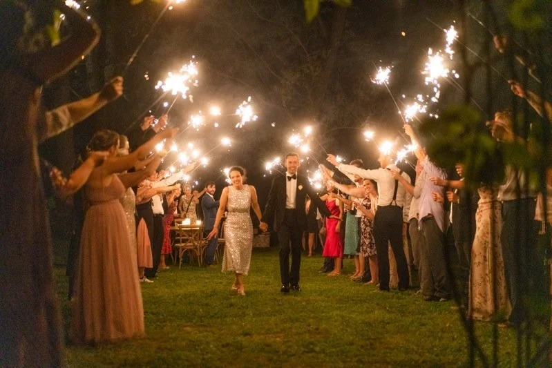 Couple walking hand in hand through a sparkler tunnel at night, surrounded by guests celebrating at an outdoor wedding reception.
