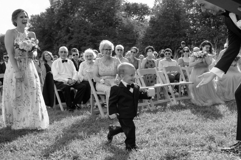 Young boy in a tuxedo running on grass at an outdoor wedding ceremony, with seated guests and bridesmaids in the background.