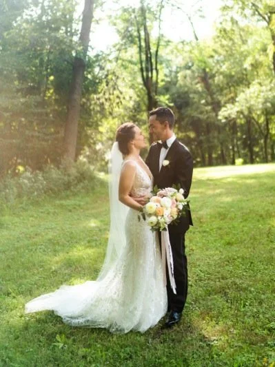 A bride and groom in wedding attire standing close together in a grassy outdoor area with trees, holding a bouquet of flowers, and gazing into each other's eyes.