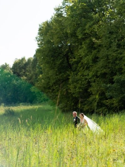 A bride and groom walking through a grassy field with large trees in the background, outdoors on a sunny day.
