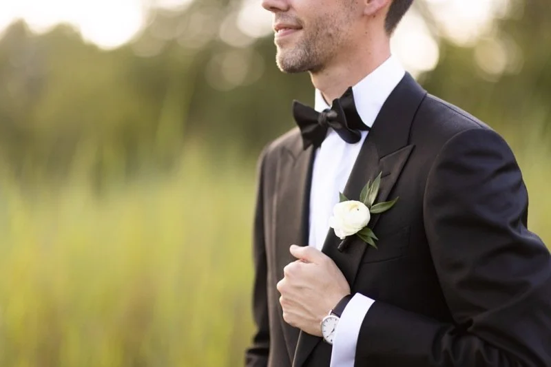 A man in a black tuxedo with a bow tie, holding a white flower boutonniere, standing outdoors.