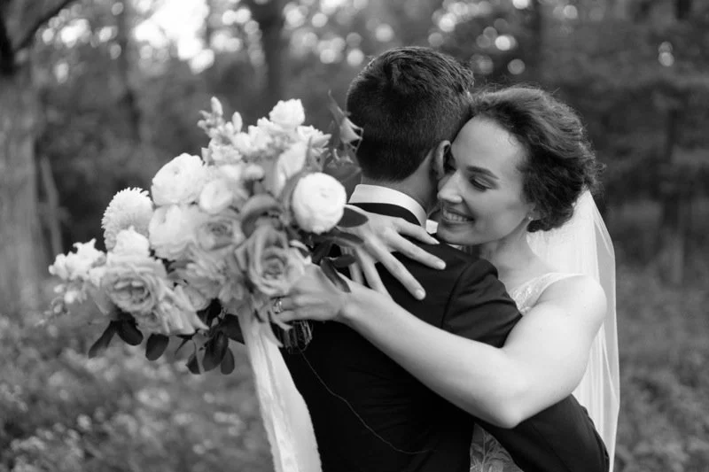 A bride and groom embracing outdoors, with the bride smiling and holding a large bouquet of flowers.