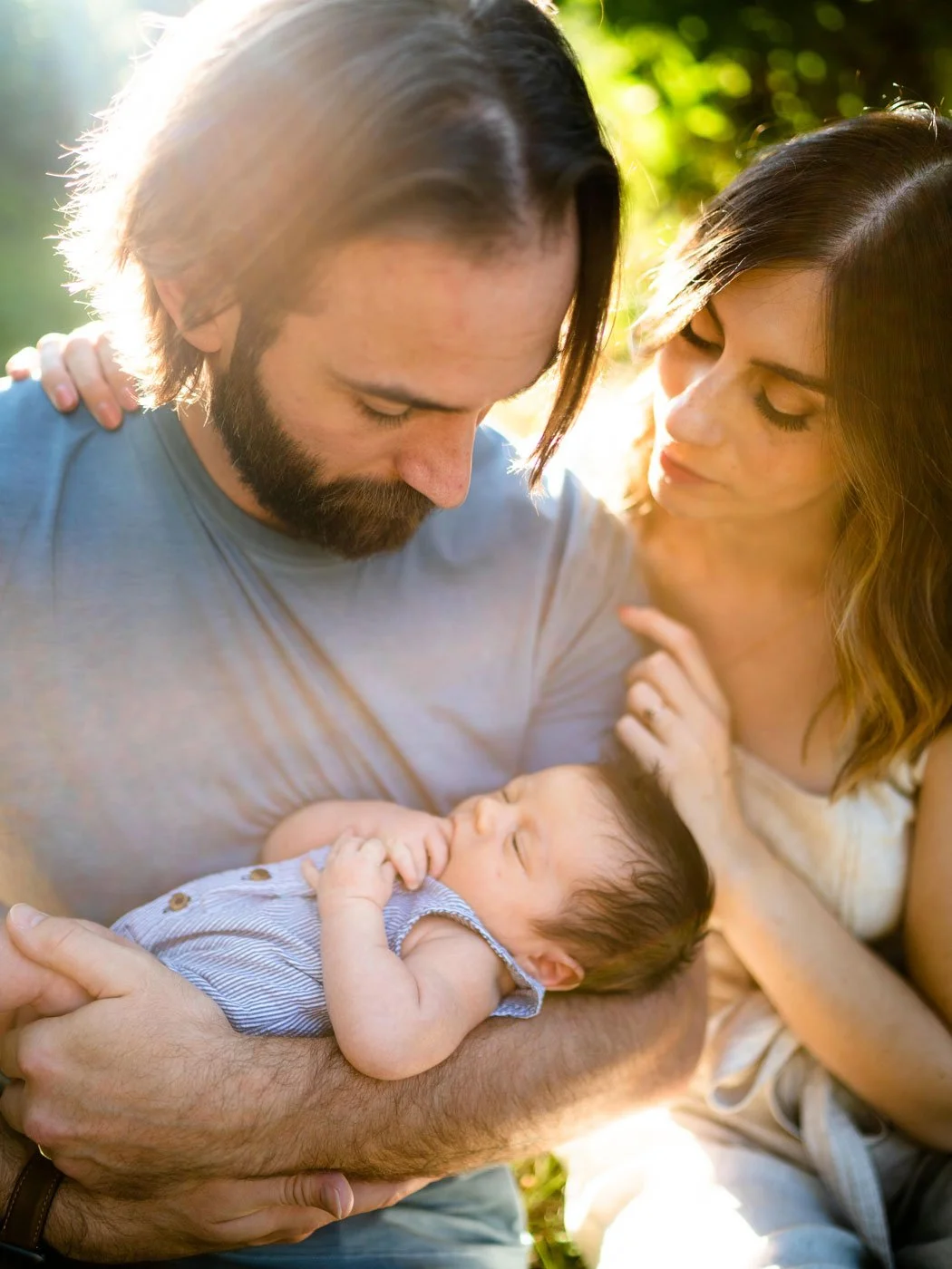 A man and woman holding a newborn baby outdoors during sunlight, with green trees in the background. The man cradles the baby in his arms while the woman gently touches the baby's head, all looking down at the baby.