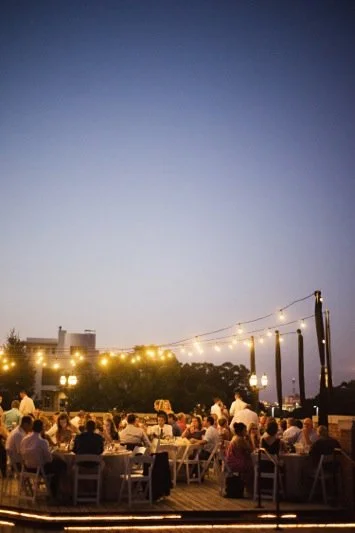 Outdoor rooftop gathering with people sitting at tables, string lights overhead, and a clear evening sky.
