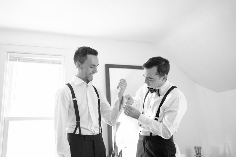 Two men in formal attire, one with suspenders and the other with a bow tie, are smiling and exchanging a fist bump in a bright room.
