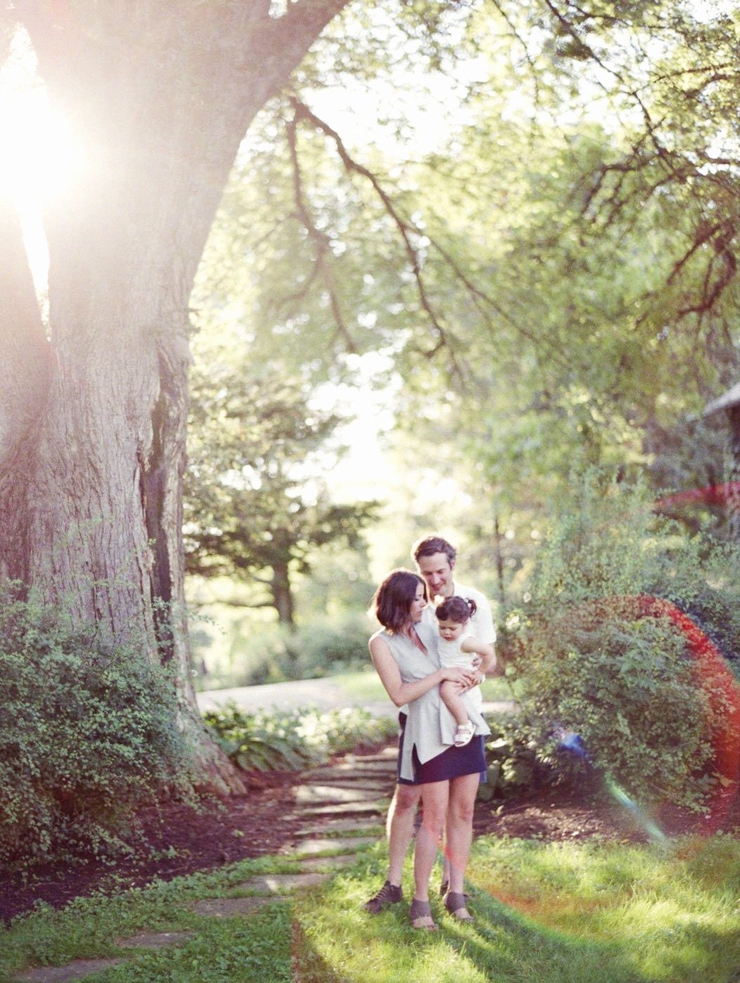 A family of three standing on a park path surrounded by trees and sunlight, with the woman holding a young girl and the man standing close behind them.