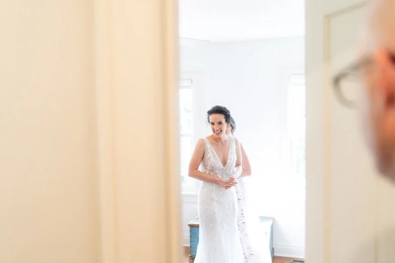 Woman in wedding dress smiling as she looks at her reflection in a mirror, with an older man partially visible in the frame.