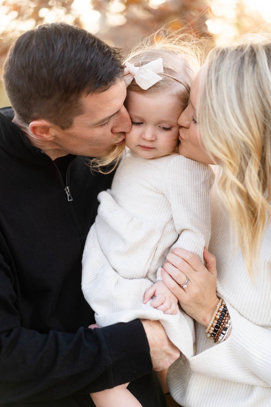A family of three sharing a tender moment, with the father and mother kissing their young daughter on the cheeks outdoors at sunset.