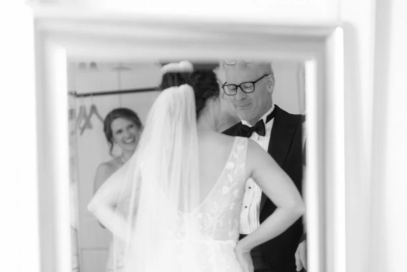 Black and white photograph of a bride in a wedding dress and veil, and a man in a tuxedo with glasses, looking at each other during a wedding ceremony, with a bridesmaid smiling in the background.