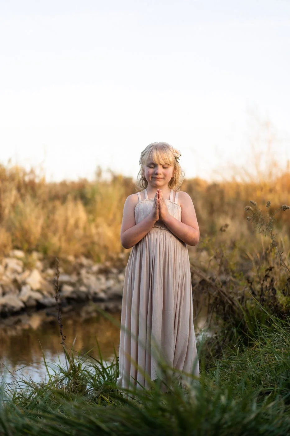 Young girl with blond hair and a flower headband wearing a beige dress, standing outdoors near a small body of water, with her hands in prayer, eyes closed, serene expression, during sunset with golden light.