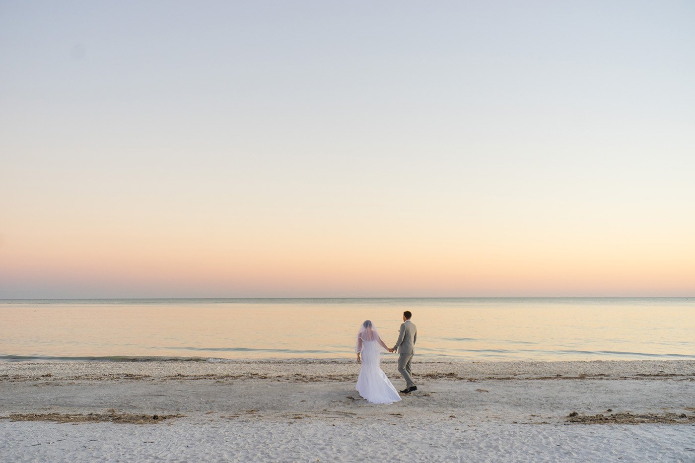 A bride and groom holding hands and walking on a beach at sunset.