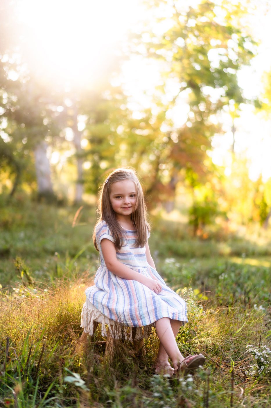 A young girl with long brown hair wearing a striped dress, sitting on a small stool outdoors in a sunlit forest or park during sunset.