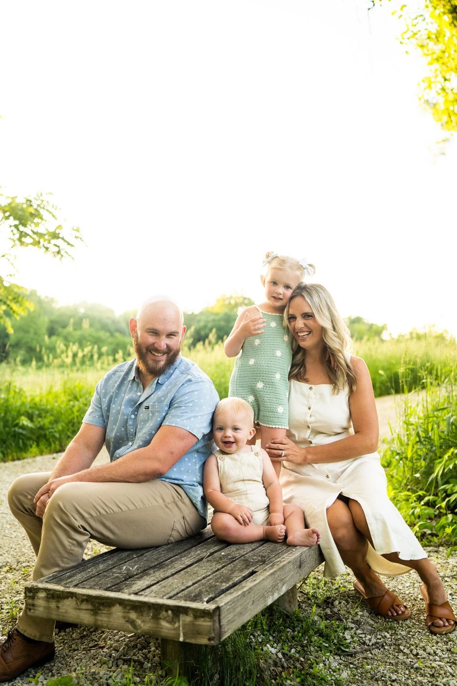 A family of four sitting on a wooden bench outdoors, with a man, woman, and two young girls, surrounded by greenery on a sunny day.