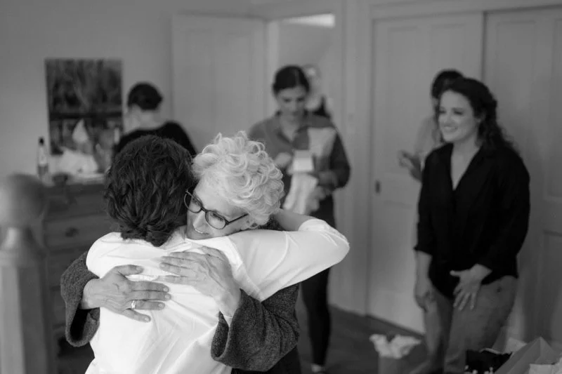 An elderly woman hugs a middle-aged woman in a heartfelt embrace, while two other women in the background smile and look at their phones in a cozy indoor setting.