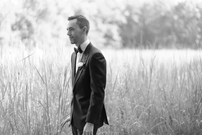 Black and white photo of a man in a tuxedo standing in a grassy field, looking to the side.