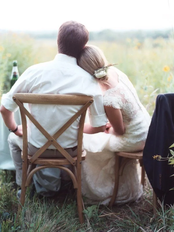 A couple sitting outdoors at a wedding or special event, embracing and looking at a scenic view, with a bottle and glass nearby.