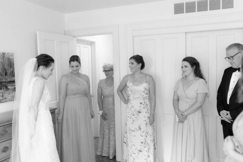 A bride in a wedding dress is bowing her head while five women and one man look at her in a room with white doors.