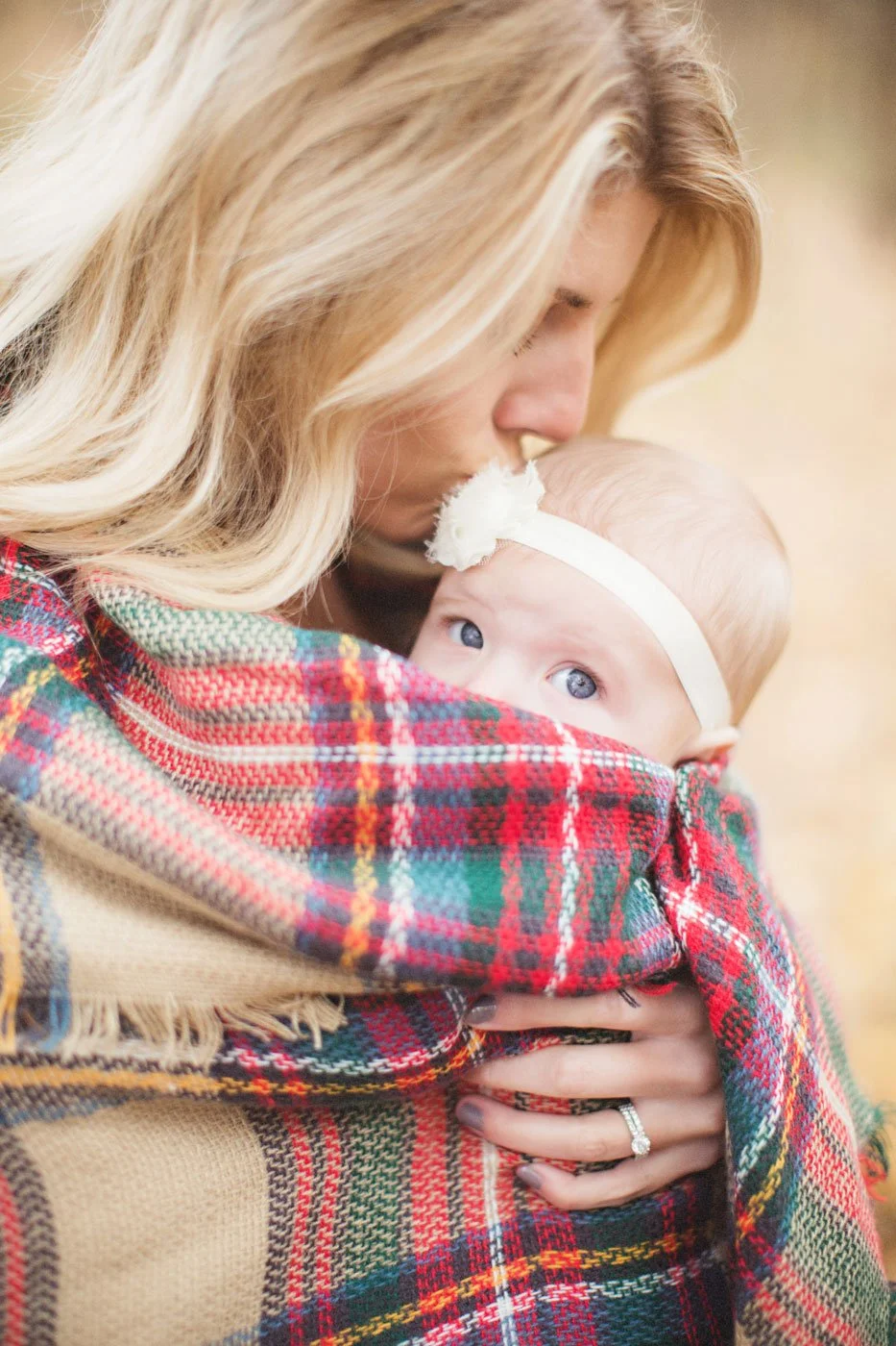 A woman with long blonde hair holding a baby wrapped in a colorful plaid blanket, with the woman wearing a matching plaid scarf while the baby has a white headband with a flower accessory.