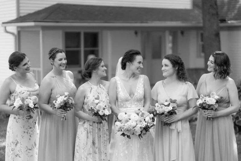 Bride and five bridesmaids standing outside in front of a house, each holding bouquets, smiling, dressed in light-colored gowns.