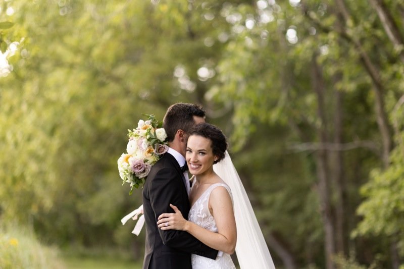 A newlywed couple smiling, with the bride wearing a white wedding dress and veil, and the groom in a black suit, standing outdoors with greenery in the background.