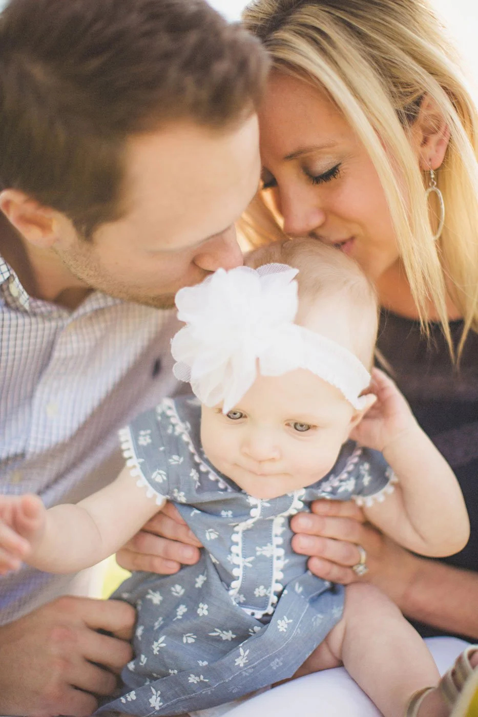 A young family, including a man and woman, hug and kiss their baby girl, who is wearing a blue floral dress and a large white headband with a bow.