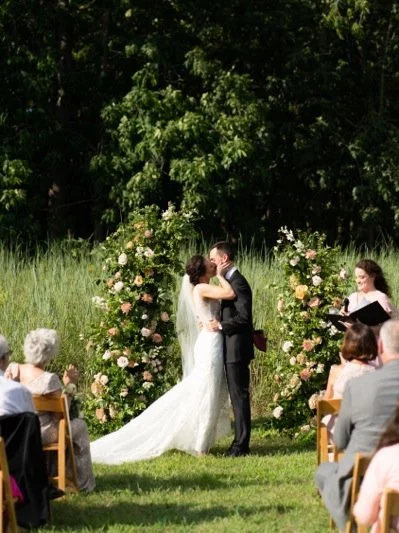 Couple getting married outdoors surrounded by flowers, officiant, and guests.